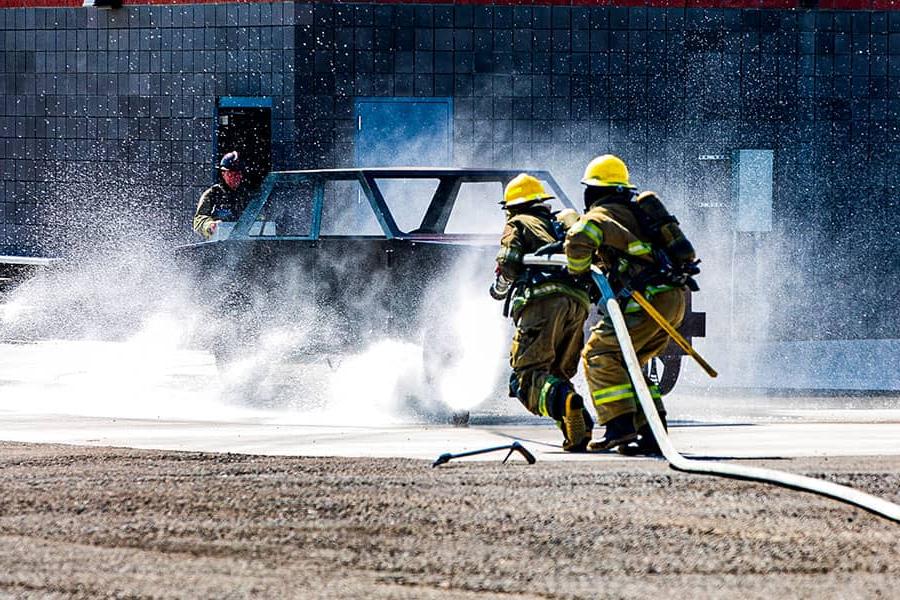 Two firefighters drag a water hose toward a burning car